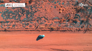 Aerial shot of a blue car driving along a red dirt track with desert plants along the side of the road. The audio description logo is in the top left corner, accompanied by the words ‘this program is audio described’. The NITV logo is in the top right hand corner. It is an abstraction of a campfire constructed from pieces of the SBS logo.