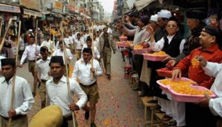 Image of Indian men walking in a ceremony, some playing instruments,  along the street being showered with petals by the crowd