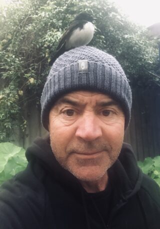 Head and shoulder photograph of Kim Scott wearing a beanie with a willy wagtail resting on his head