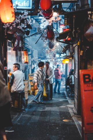 three people in a bustling alleyway in Japan with lanterns and cherry blossom hanging above