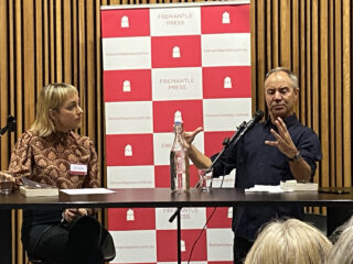 Kim Scott and Molly Schmidt sitting at a table, with microphones and a red and white banner background, discussing the book