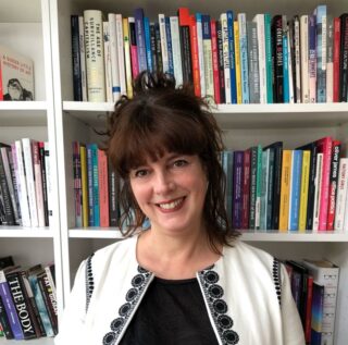 Head and shoulders image of Rosalind Gill smiling sitting in front of a book library wearing a black and white top