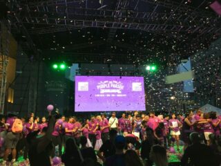 Image of the finale of the Purple Parade 2019. It features a group of people standing on stage dressed in purple, while confetti sprinkles onto them. In the foreground you can see the silhouettes of other audience members. 