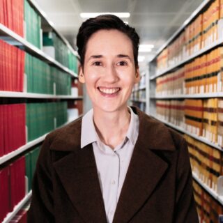 Head and shoulders image of Emilia smiling, standing in an aisle of books at a library