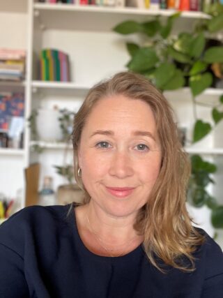 Head and shoulders photo of Kim Cousins smiling sitting in front of a white bookcase with books and plants growing on it.,